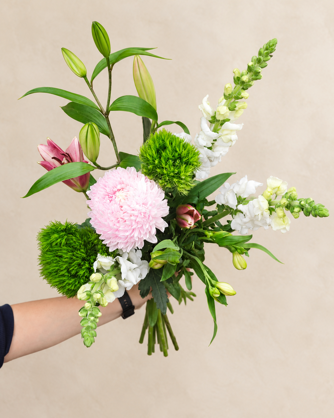 Hand-held Petite bouquet with a pink chrysanthemum, white snapdragons, lily buds and green foliage against an ivory background.
